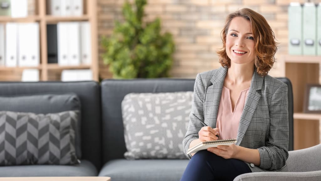 A woman sits on a couch in an office, smiling and holding a notepad and pen. Bookshelves filled with insightful reads and potted plants are in the background, subtly hinting at her curiosity about topics like "How Much Does BetterHelp Pay Therapists.